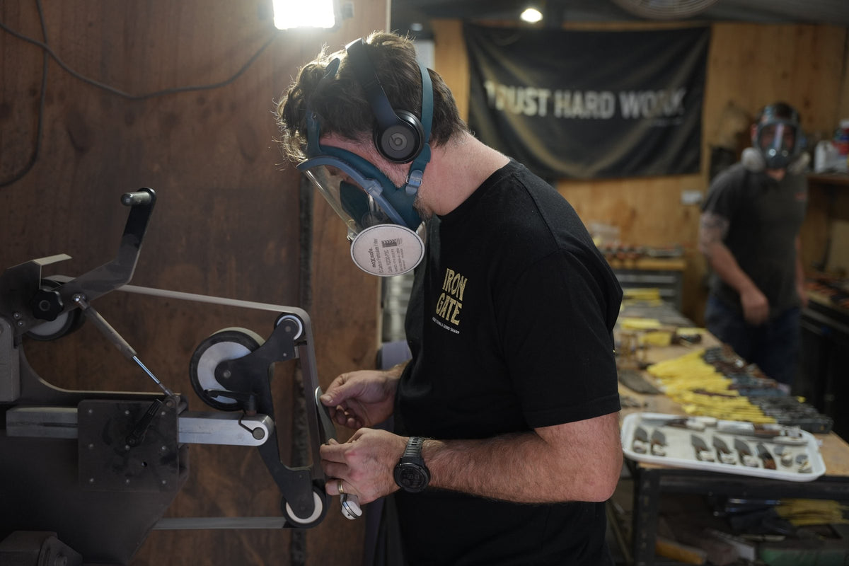Knifemaker in a respirator grinding a blade under the Trust Hard Work banner