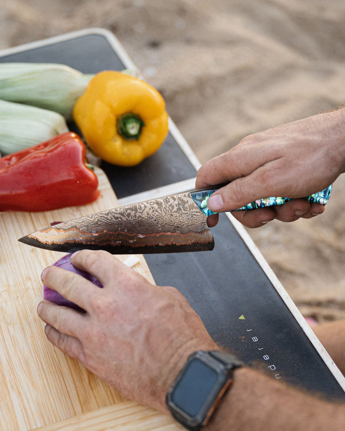 Hands slicing red capsicum on a board with a damascus chef's tool, fresh vegetables alongside