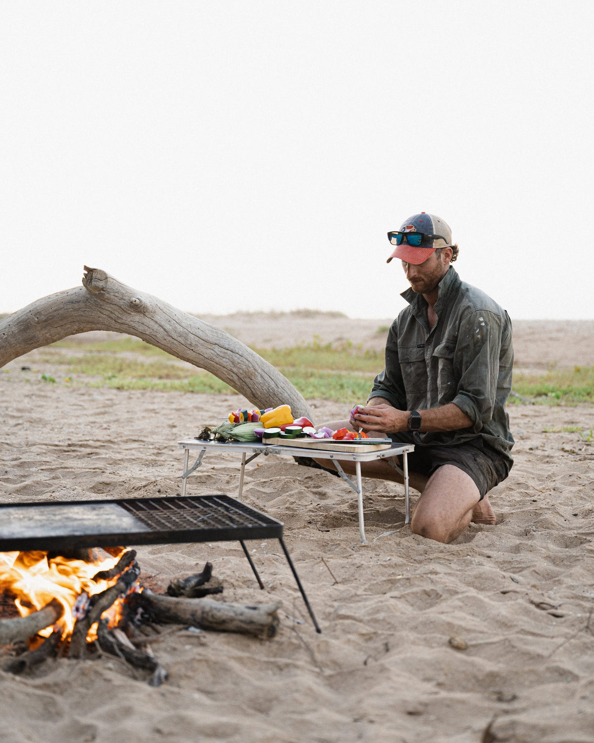 A man prepares food at a small camp table on the beach beside a crackling campfire and driftwood