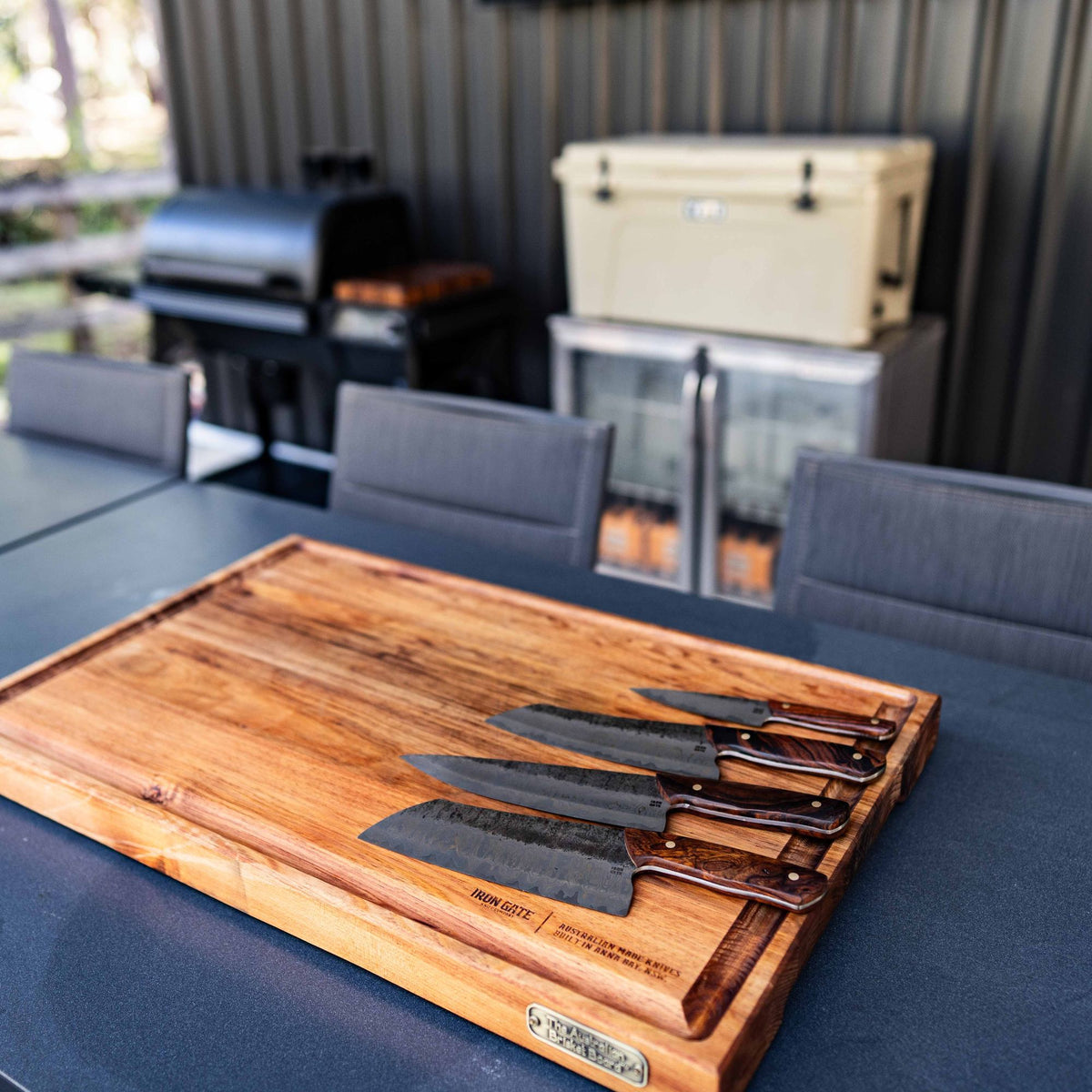 A kitchen set laid on a large timber board on an outdoor dining table, grill and cooler behind