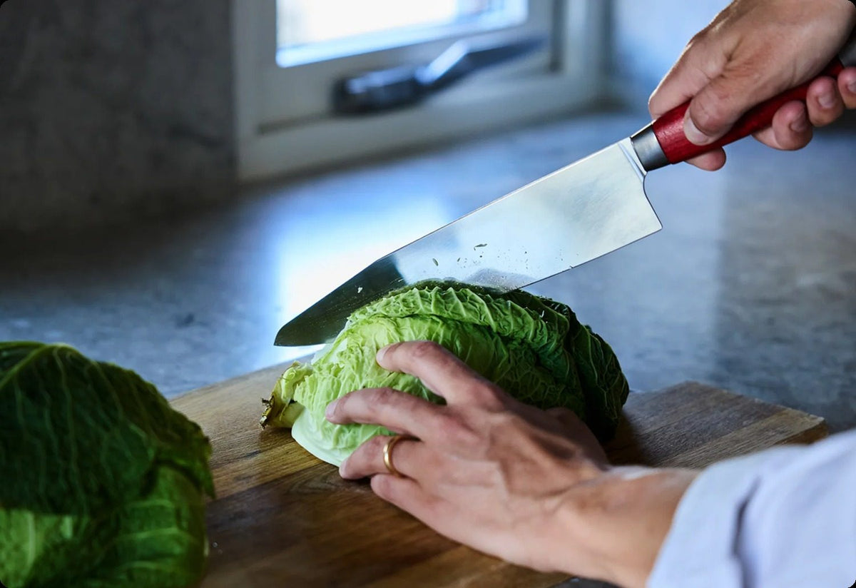 A hand slicing a cabbage on a kitchen bench with a red-handled chef's blade