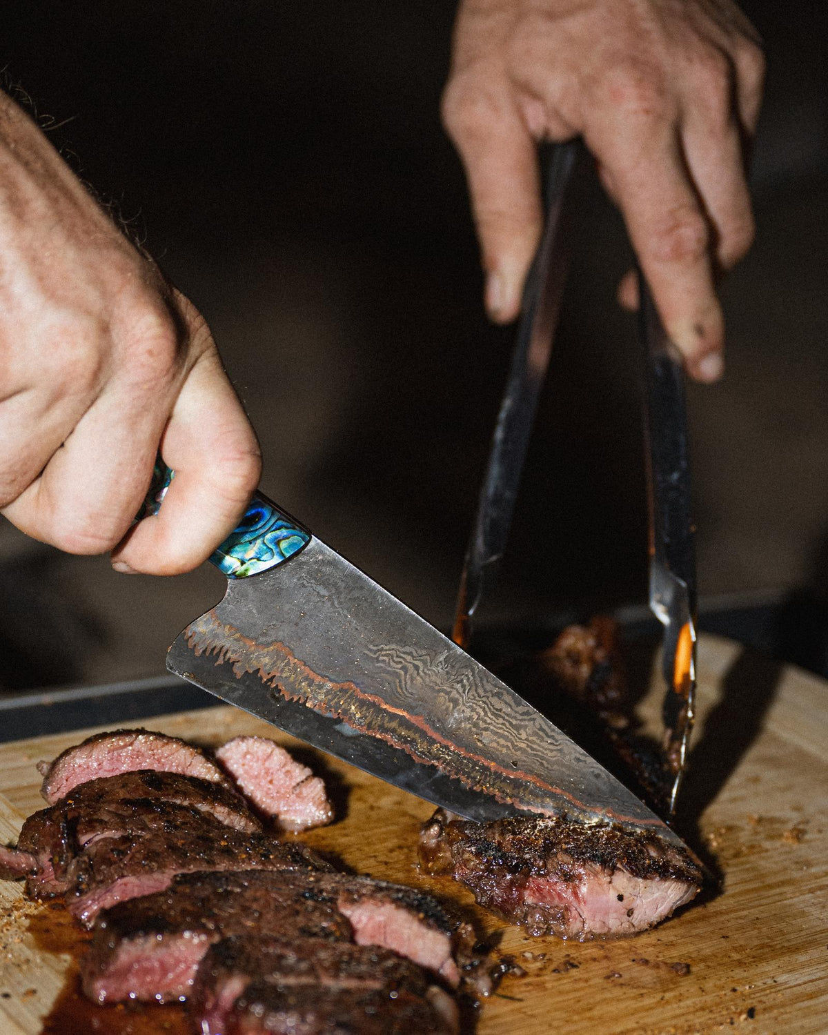 Hands carving a cooked steak on a board with an abalone-handled chef's blade