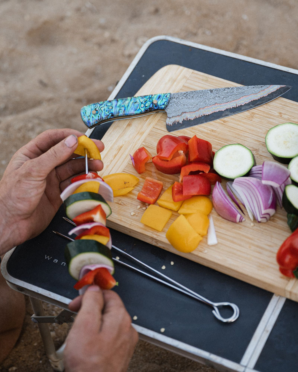 Hands threading vegetable skewers beside a board of chopped capsicum, zucchini and onion