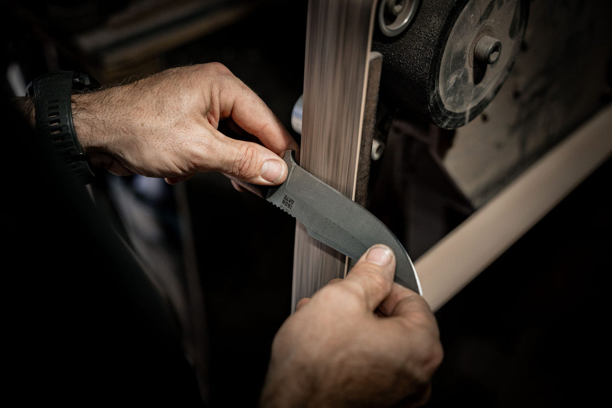 Two hands hold a bare steel blank flat to a grinding belt in low light