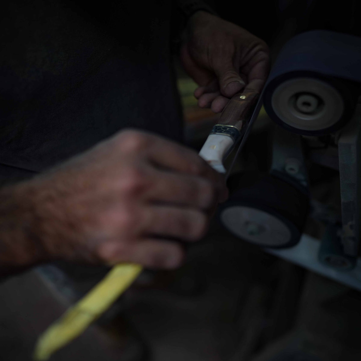 Hands sanding a handle on a small sanding wheel in a dim workshop